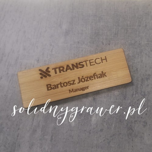 prostokątny identyfikator osobowy wykonany z naturalnego bambusa o wymiarach 75×25 mm. Na powierzchni znajduje się ciemny grawer laserowy przedstawiający dane użytkownika.